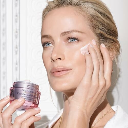 Woman applying face cream from Estee Lauder jar.